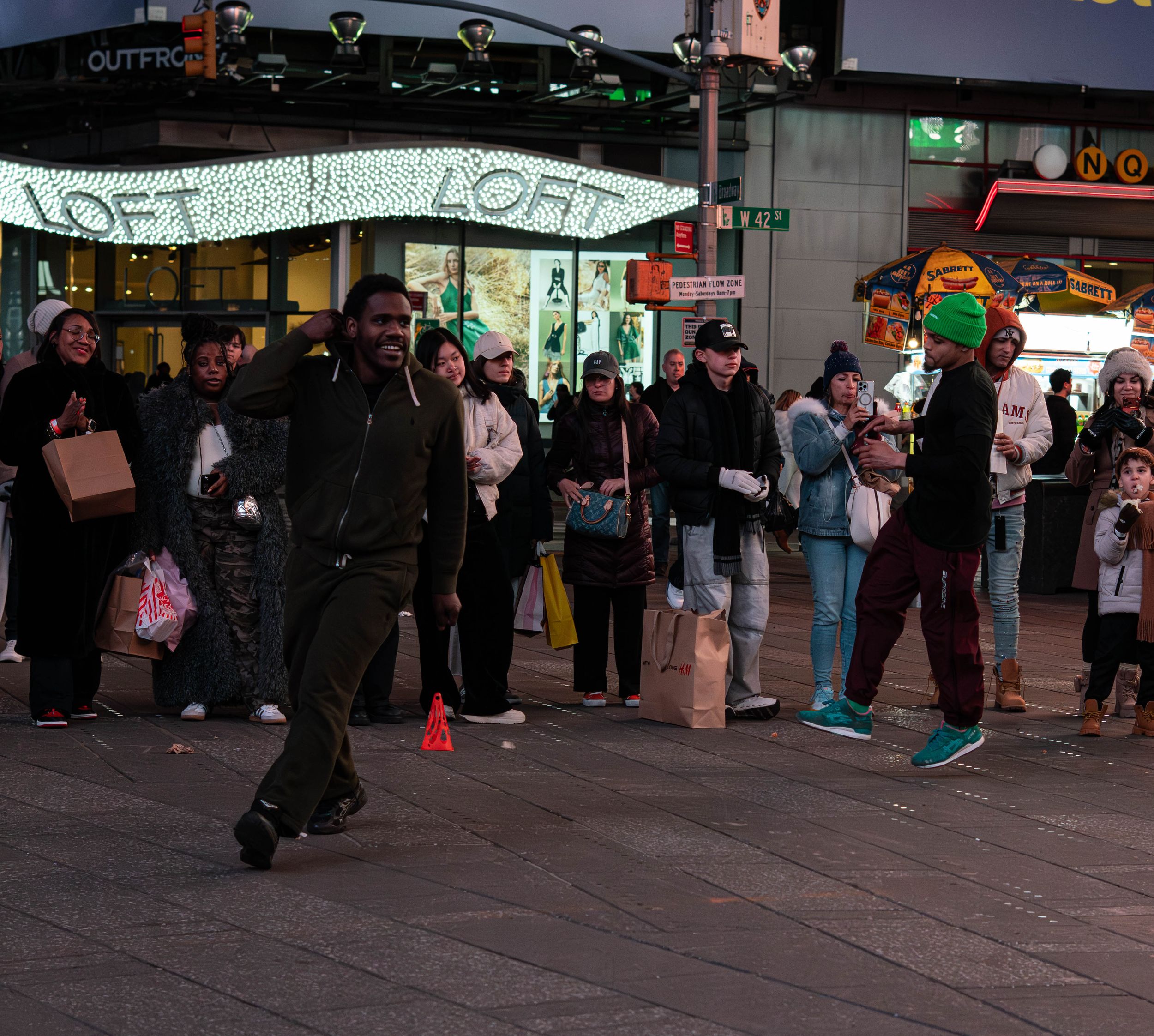 People dancing and watching street performers at night near a LOFT store in a busy urban shopping district.