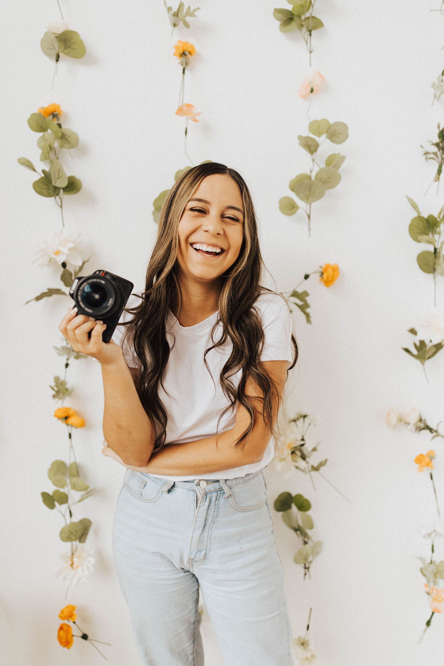 Smiling woman holding a camera in front of a white wall decorated with floral garlands.