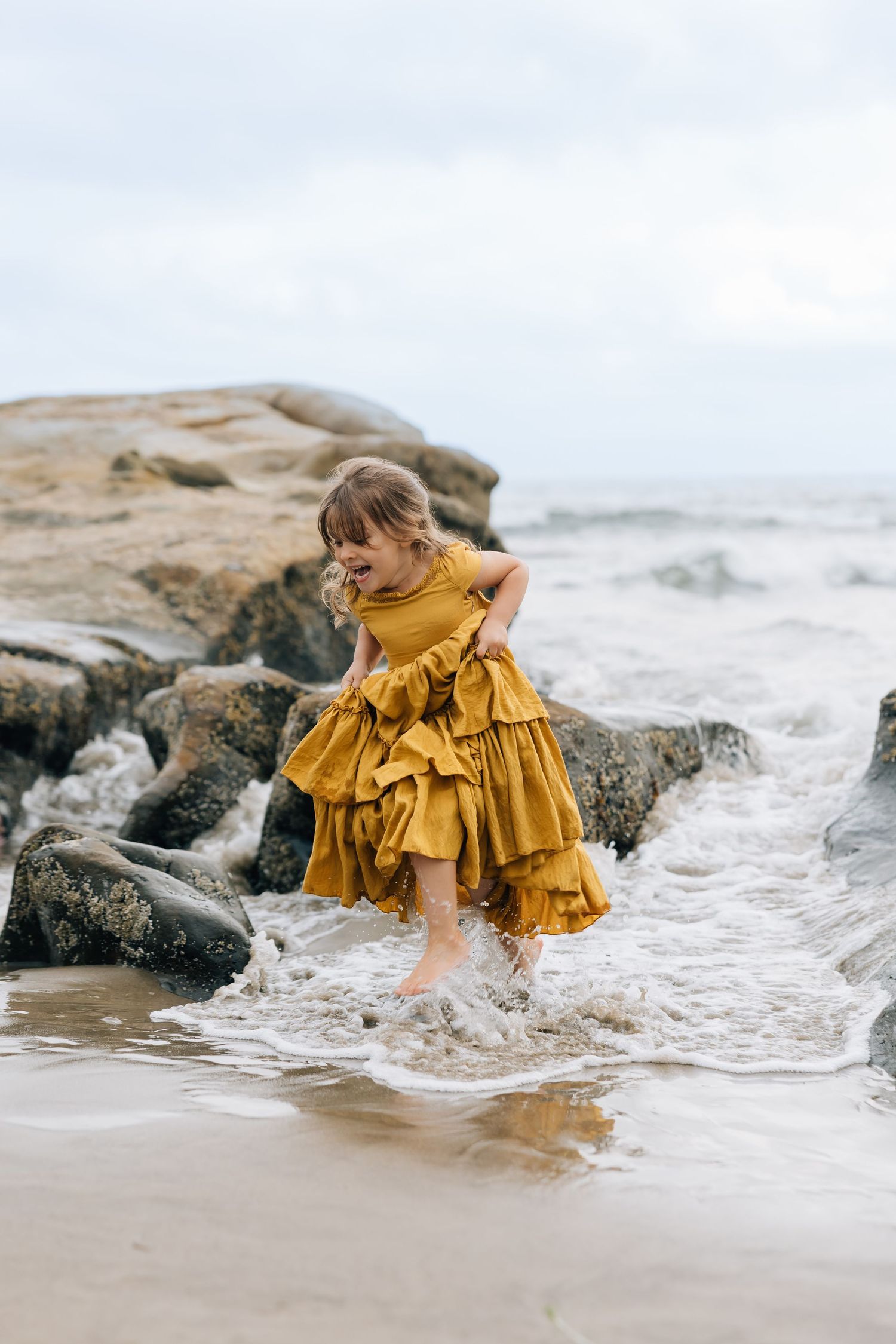 A young girl in a golden ruffled dress plays in ocean waves on a rocky beach.