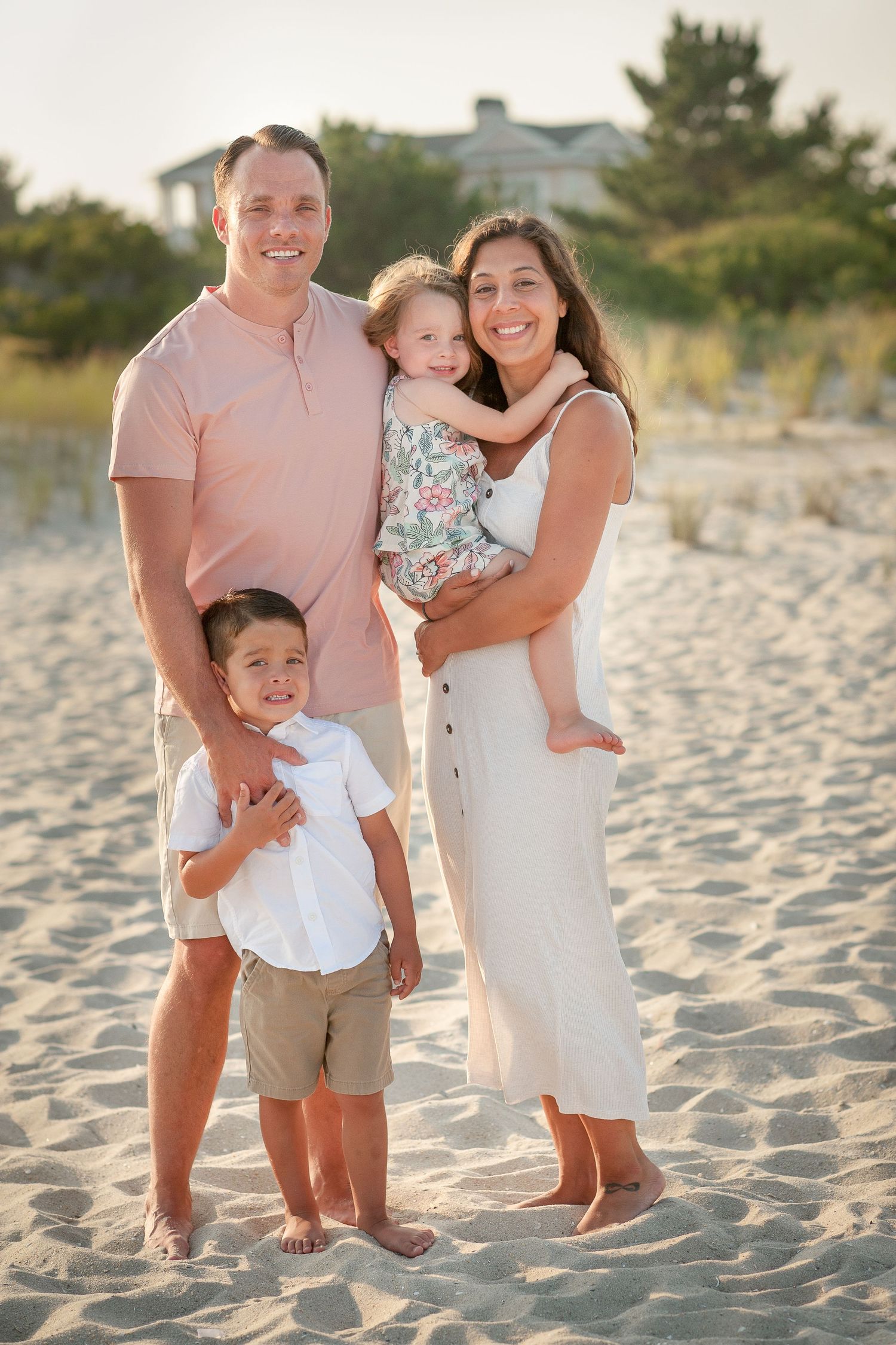 Family of four smiling together on a sandy beach, parents holding young children in warm golden light.