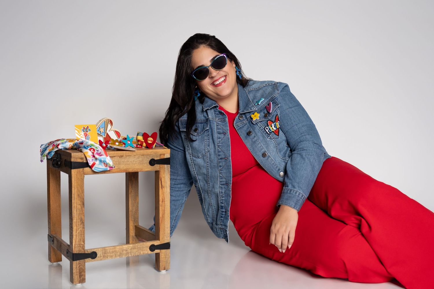 Woman in red dress and denim jacket with patches, wearing sunglasses, posing beside a wooden table with colorful items.