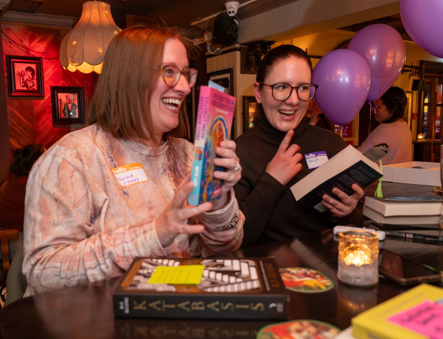 Two women laugh while holding books at a book club event with purple balloons and candles in background.