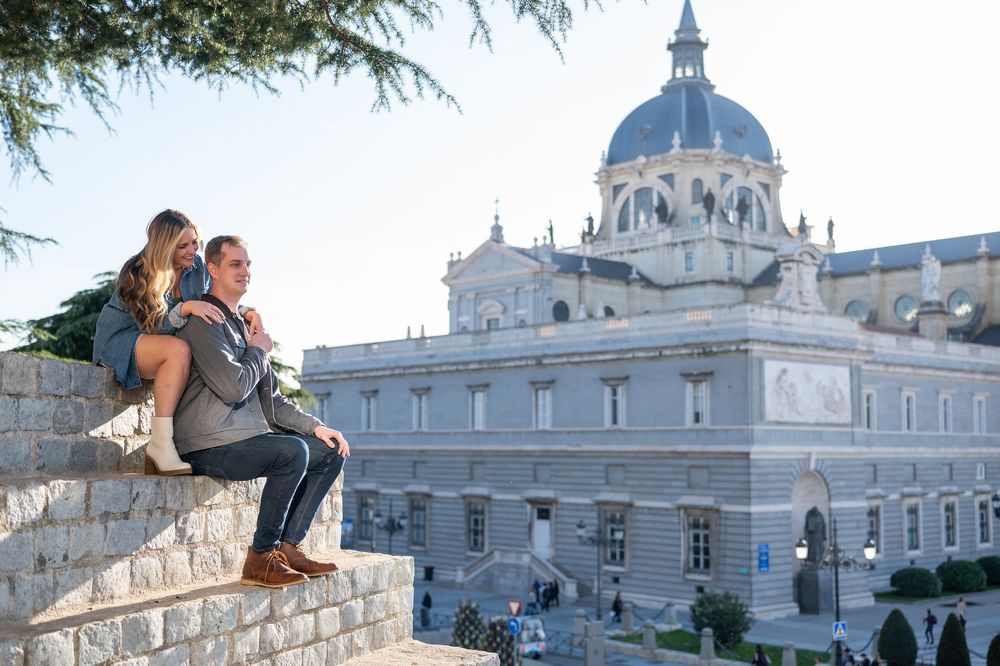 Couple sitting on stone wall with Almudena Cathedral in Madrid, Spain in the background on a sunny day.