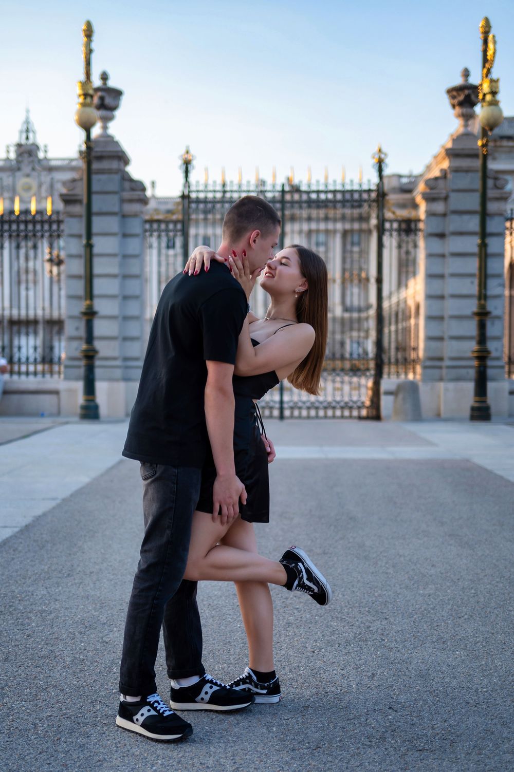Couple sharing a romantic kiss in front of an ornate palace gate, both dressed in black outfits.
