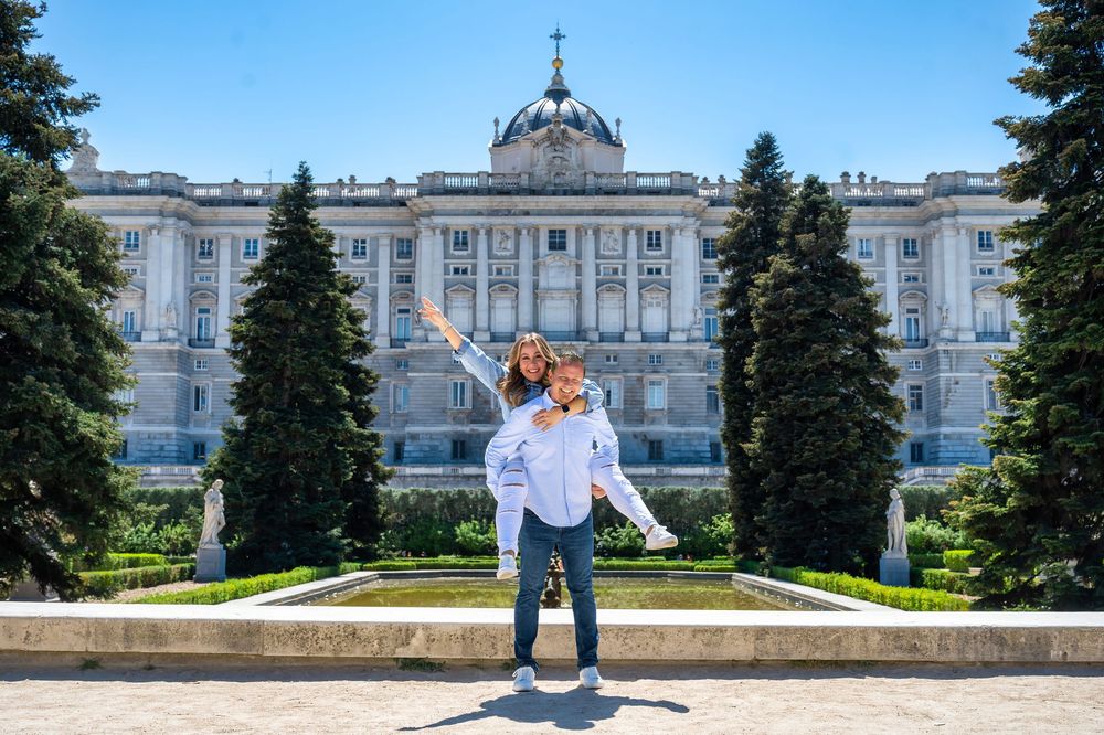 Couple posing playfully in front of the Royal Palace of Madrid surrounded by lush green trees on a sunny day.