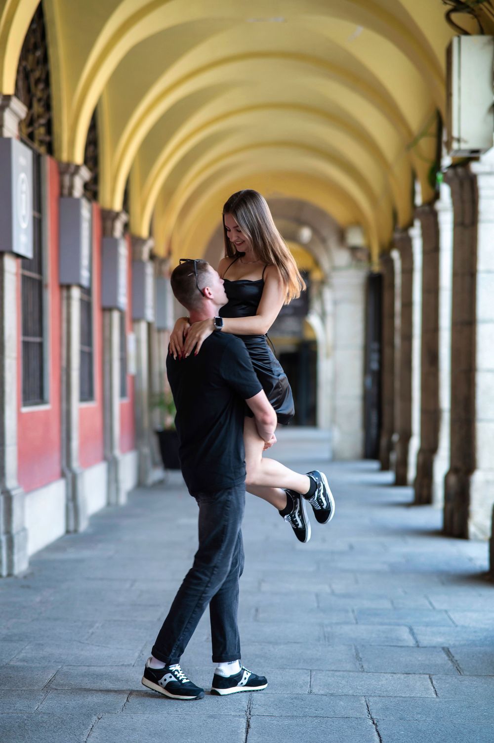 A couple in black outfits share a romantic moment under arched arcade, man lifting woman in European city.
