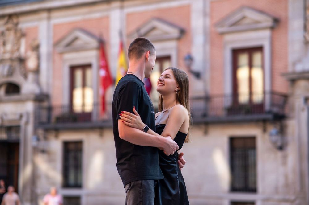 Romantic couple embracing in front of a historic building in Madrid, Spain, with a flag in the background.