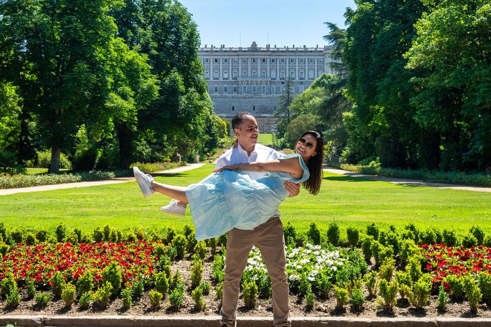 Couple posing in Madrid's Campo del Moro gardens, man lifting woman in blue dress with Royal Palace in background.