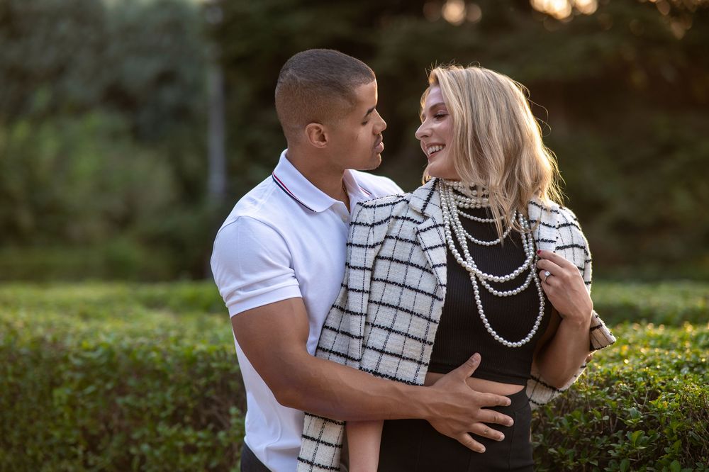 Couple embracing outdoors, woman in pearl necklace and checkered top, man in white polo, green hedges background.