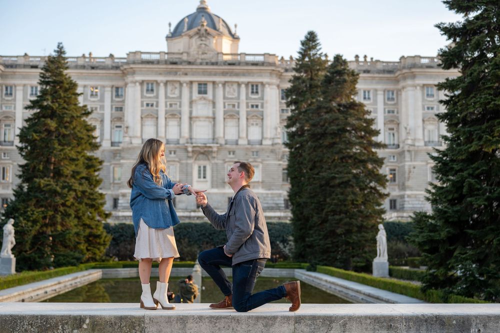 Man proposes to woman on one knee at the Royal Palace of Madrid, Spain, surrounded by lush trees and fountains.