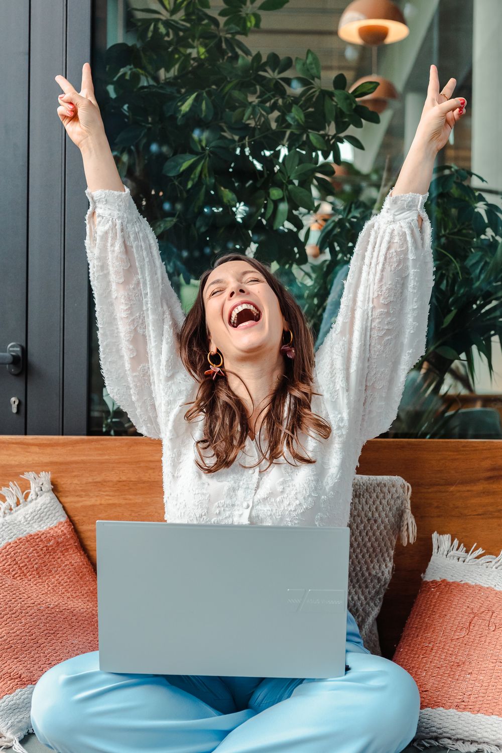 Excited young woman raising arms in victory gesture with laptop on lap, sitting on couch surrounded by green plants.