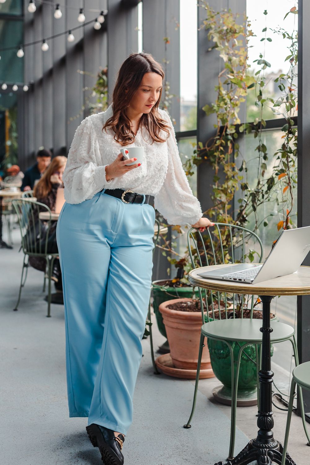 Woman in white blouse and light blue wide-leg pants holds coffee cup while working on laptop at outdoor cafe.
