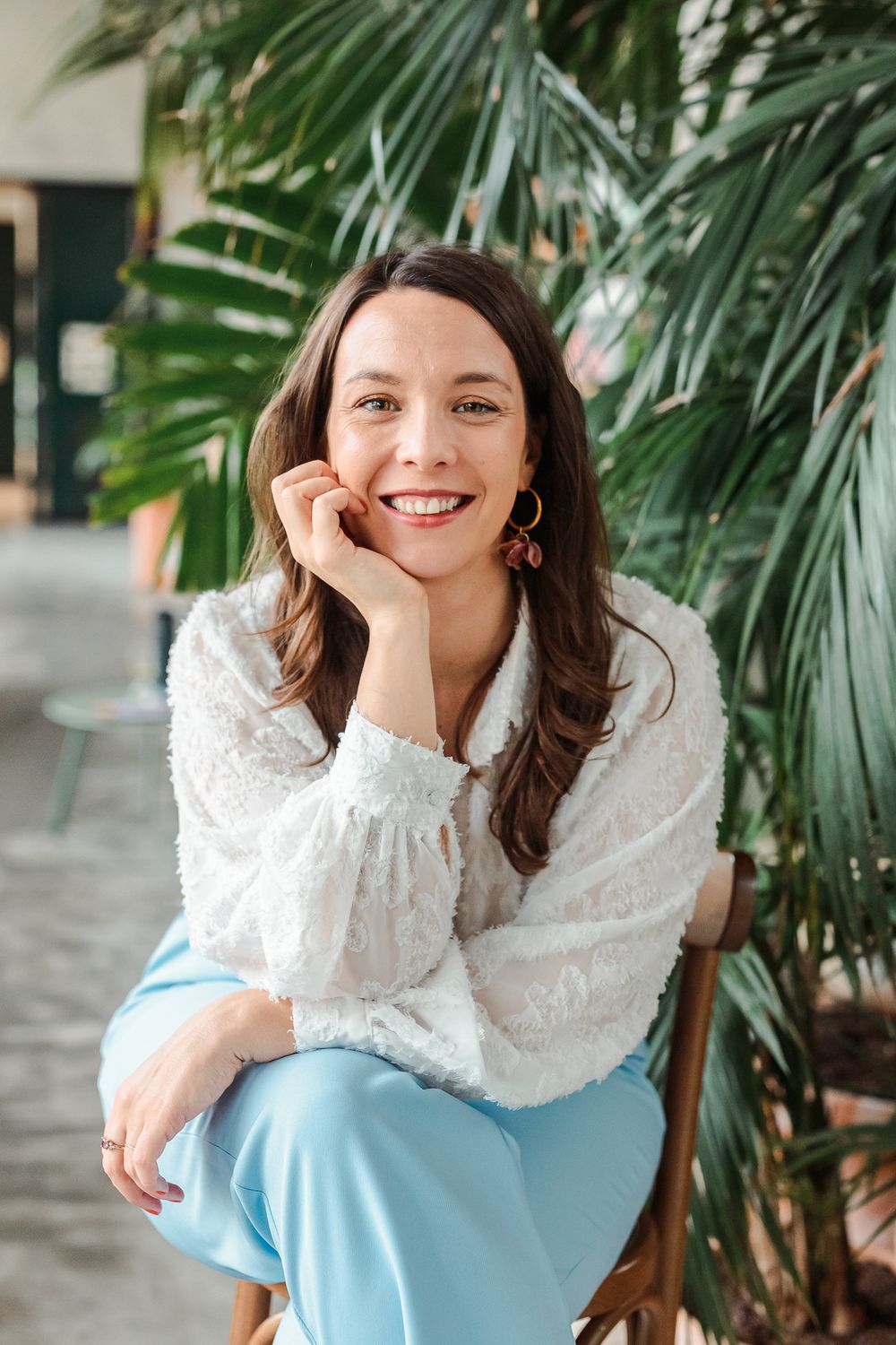 Smiling brunette woman in white lace blouse and blue pants sitting in chair with tropical plants behind her.