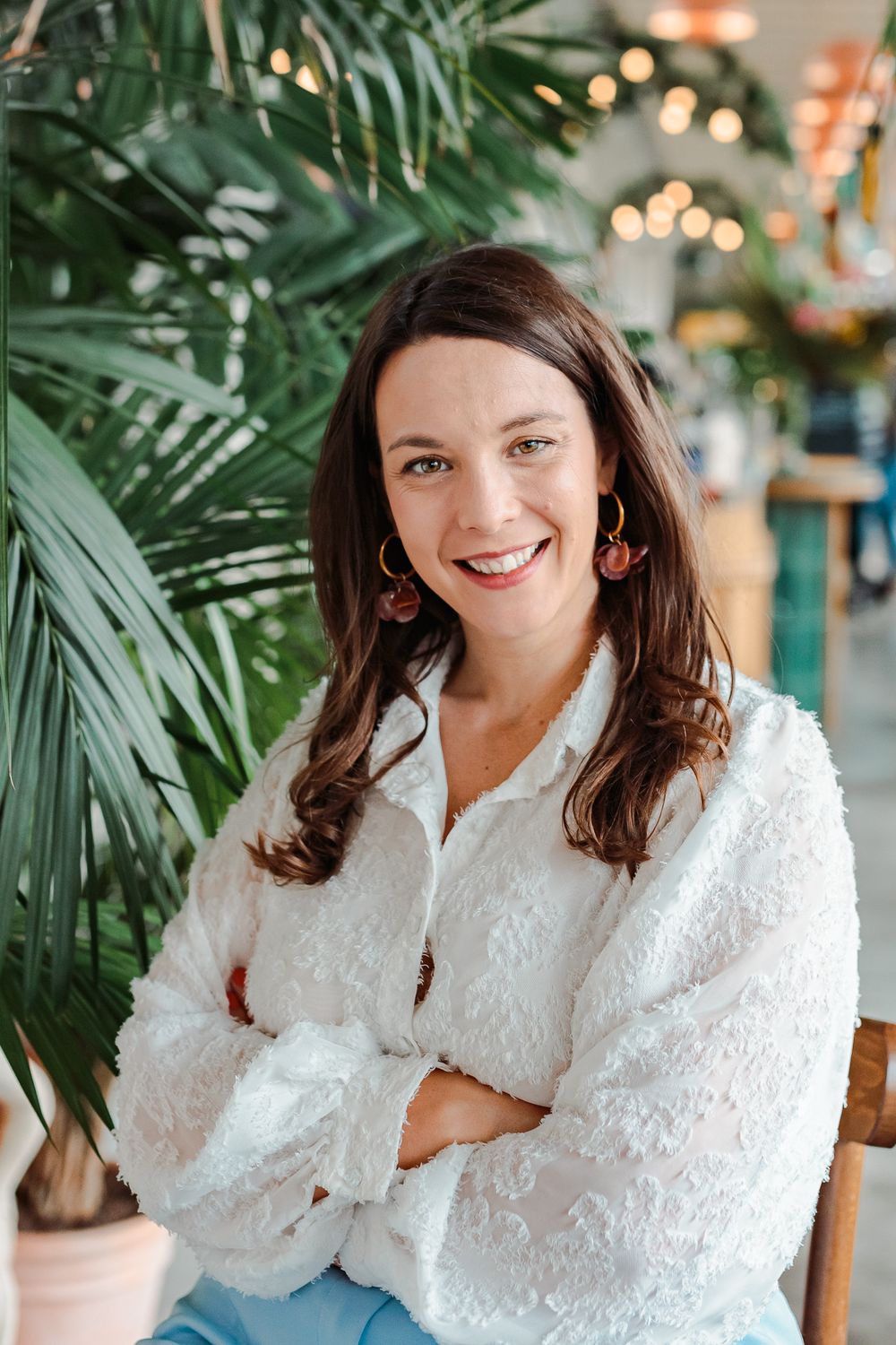 Smiling woman with dark hair, white textured blouse, crossed arms, seated near tropical plants in bright cafe.