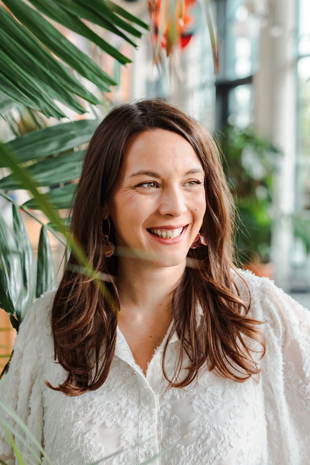 Smiling brunette woman in white lace blouse surrounded by tropical plants in bright indoor setting.