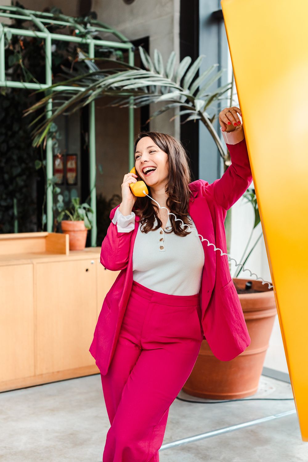 Happy woman in pink suit laughing while talking on retro yellow phone in modern interior.
