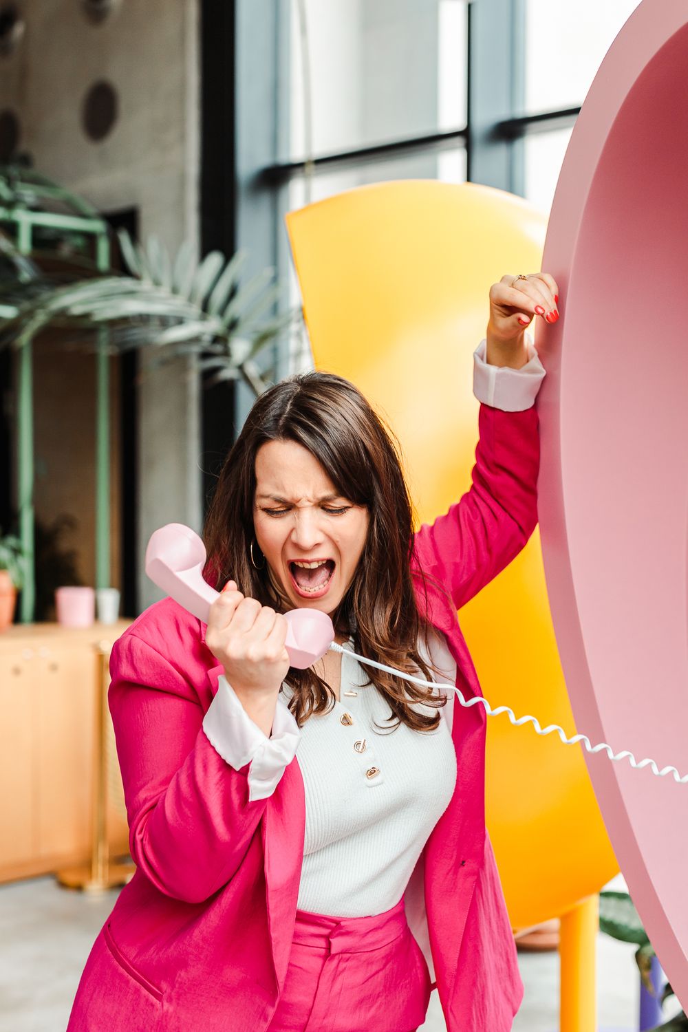 Angry woman in pink blazer screaming at pink telephone handset in colorful office space.