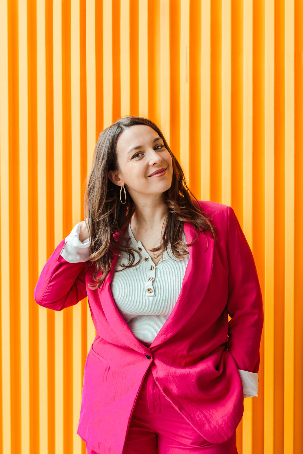 Woman in pink blazer suit smiling against bright orange striped wall.