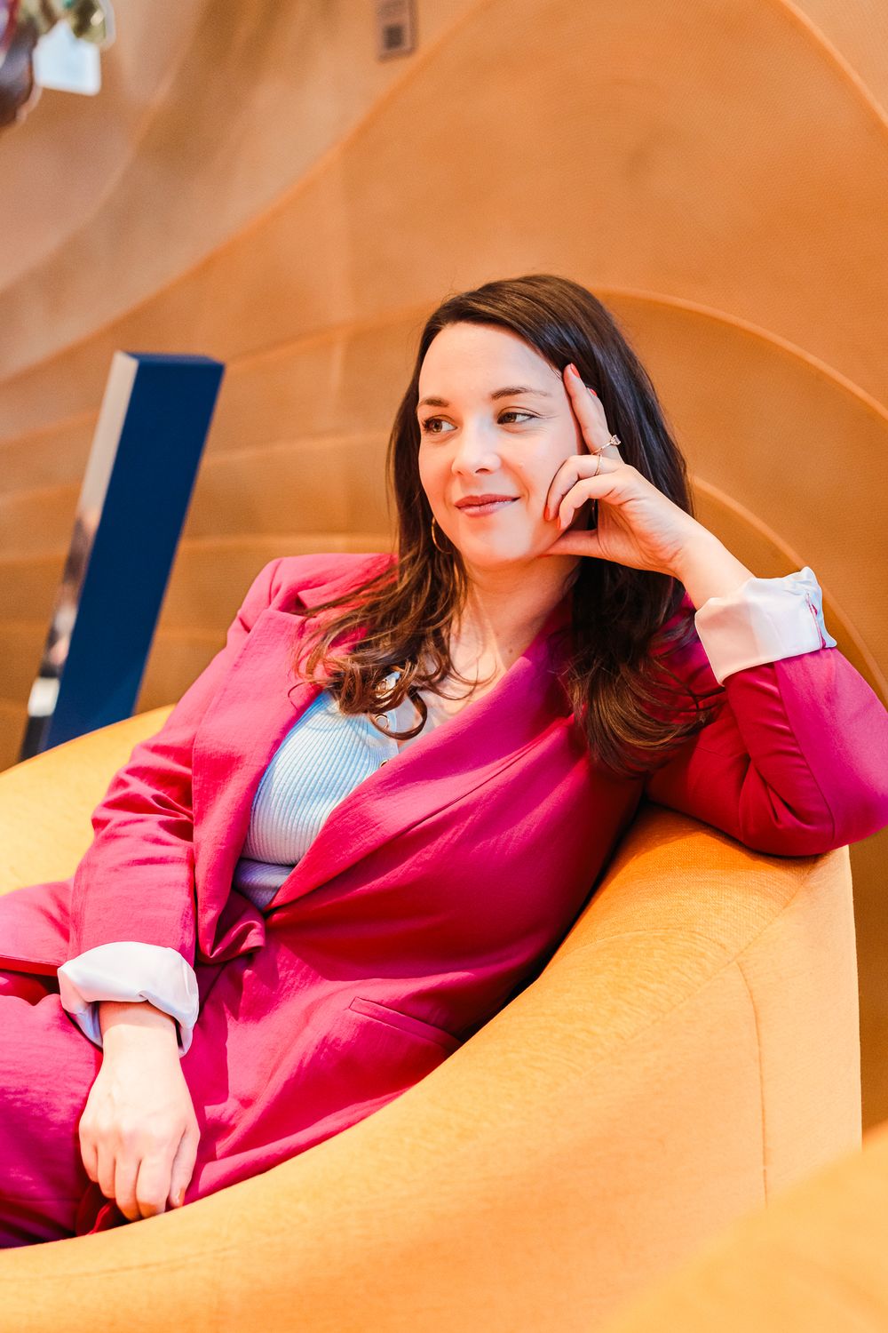 Brunette woman in pink suit relaxing in yellow chair with curved wooden architecture in background.