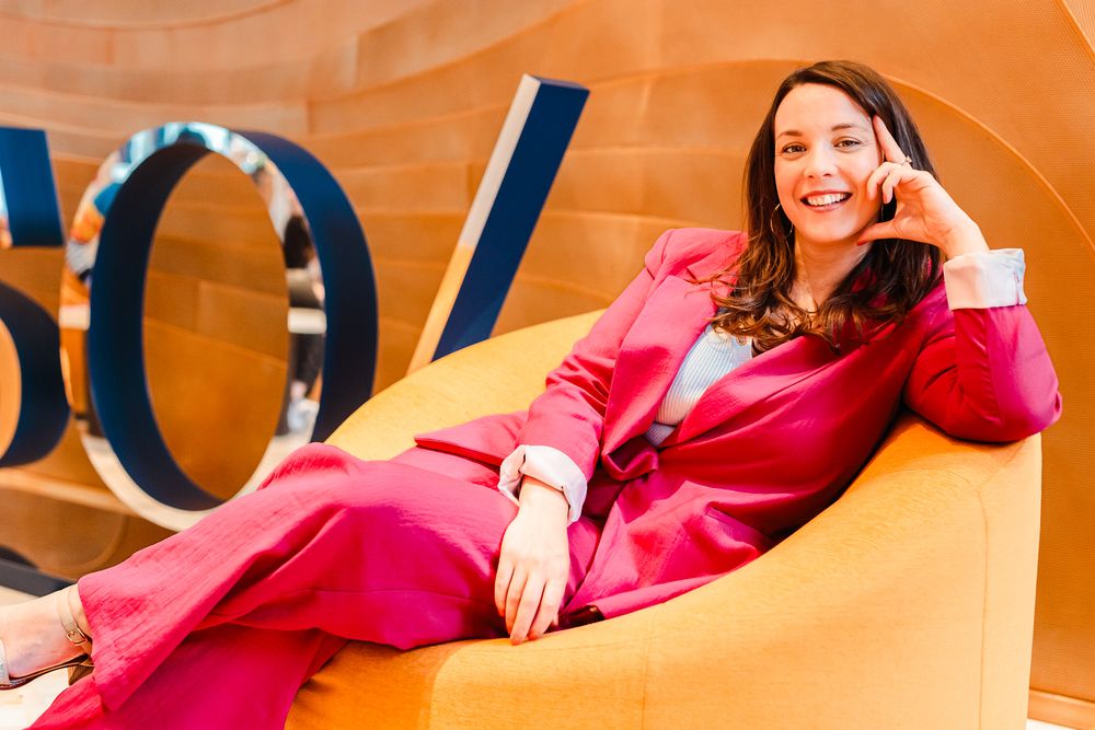 Woman in pink suit relaxing in yellow chair, smiling confidently in front of decorative gold backdrop.