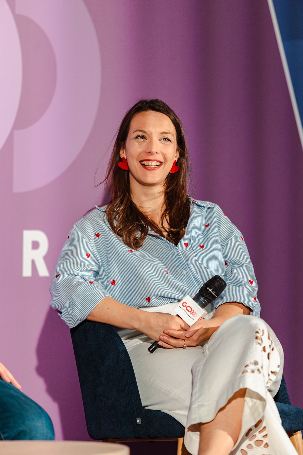 Smiling woman with red earrings holding microphone at panel event, wearing blue heart-print blouse on purple stage backdrop.