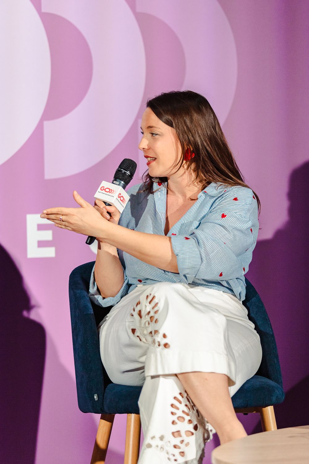 Woman in blue shirt and white pants speaks into microphone while seated on stage at an event.
