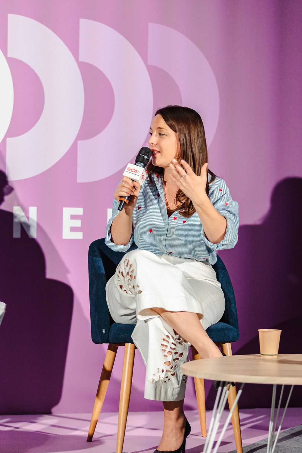 Brunette woman in blue blouse and white pants gestures while speaking at a pink-themed conference.