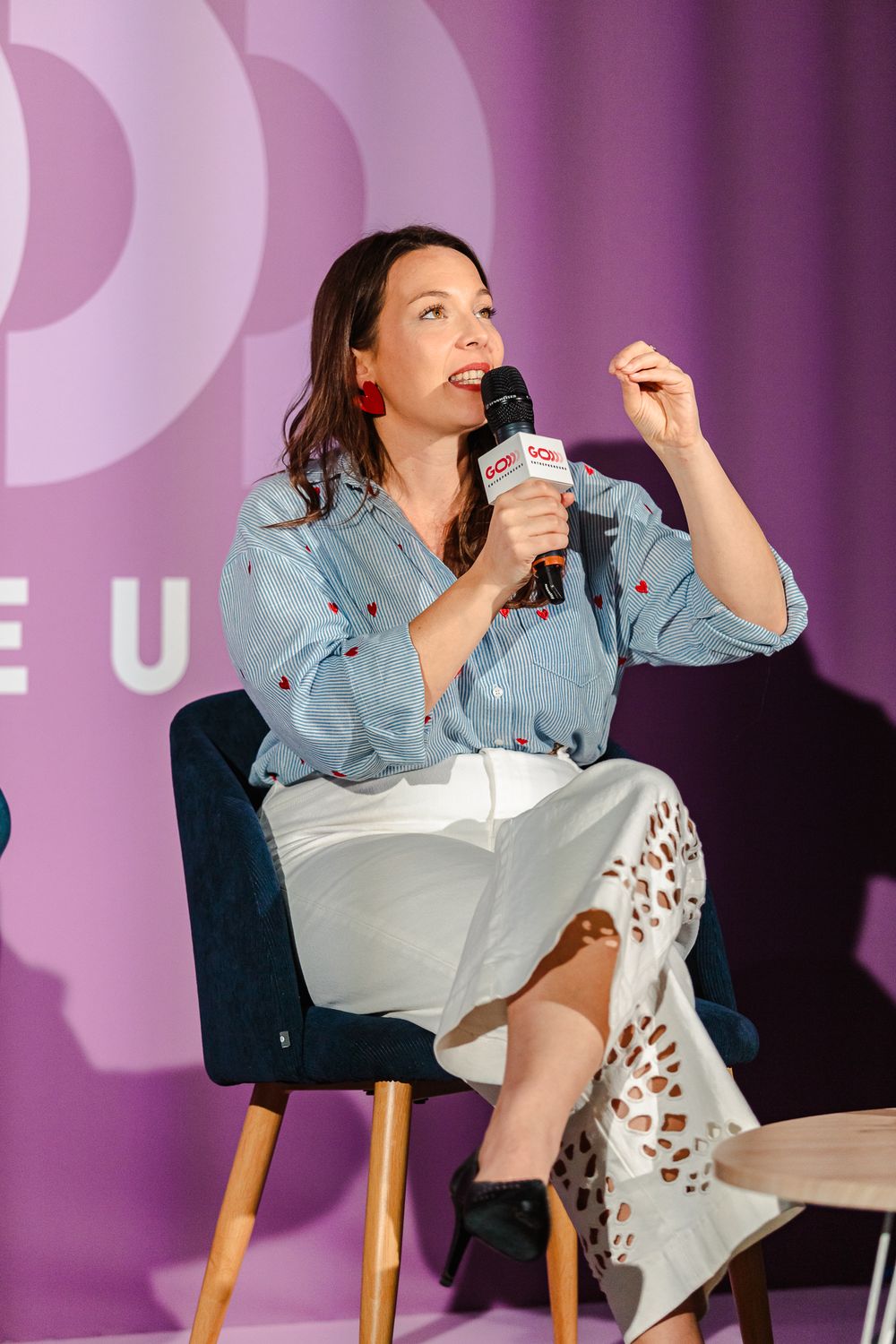 Woman in blue shirt speaks into microphone while seated on stage at a conference event.