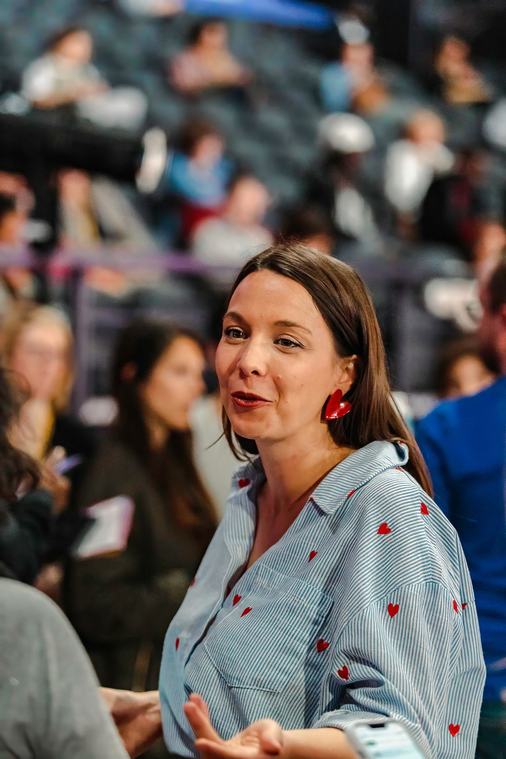 Woman with red heart earrings and heart-print blue shirt sits in a crowded auditorium, gesturing while speaking.