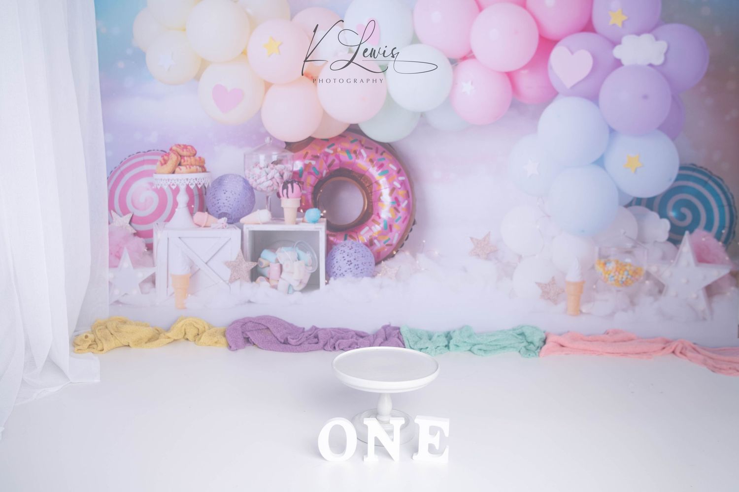 Pastel balloon arch backdrop with rainbow, donuts, and ONE sign for a first birthday cake smash setup.