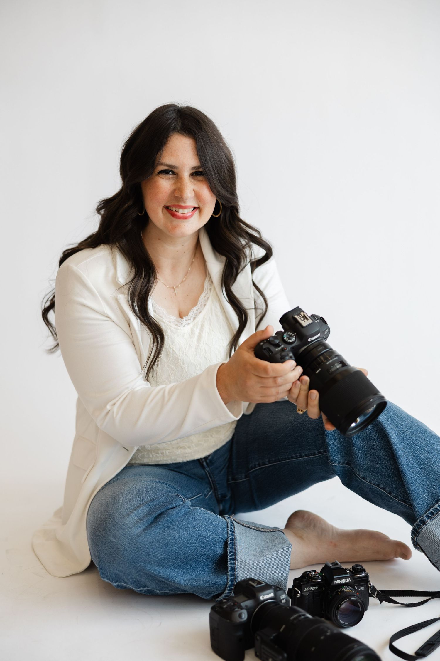 Smiling female photographer sitting on stool holding DSLR camera, wearing white blazer and jeans against white background.