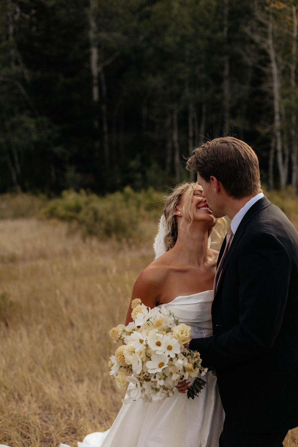 Bride and groom share a romantic moment in an autumn field, bride holding white floral bouquet in wedding dress.