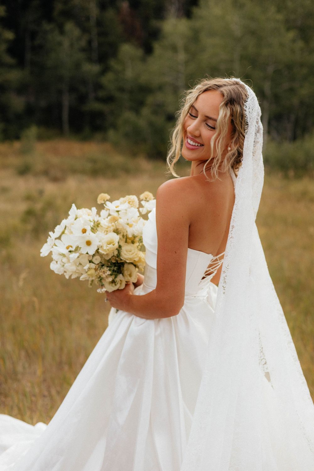 Smiling bride in strapless white gown holding white bouquet with veil, standing in golden meadow with trees behind.