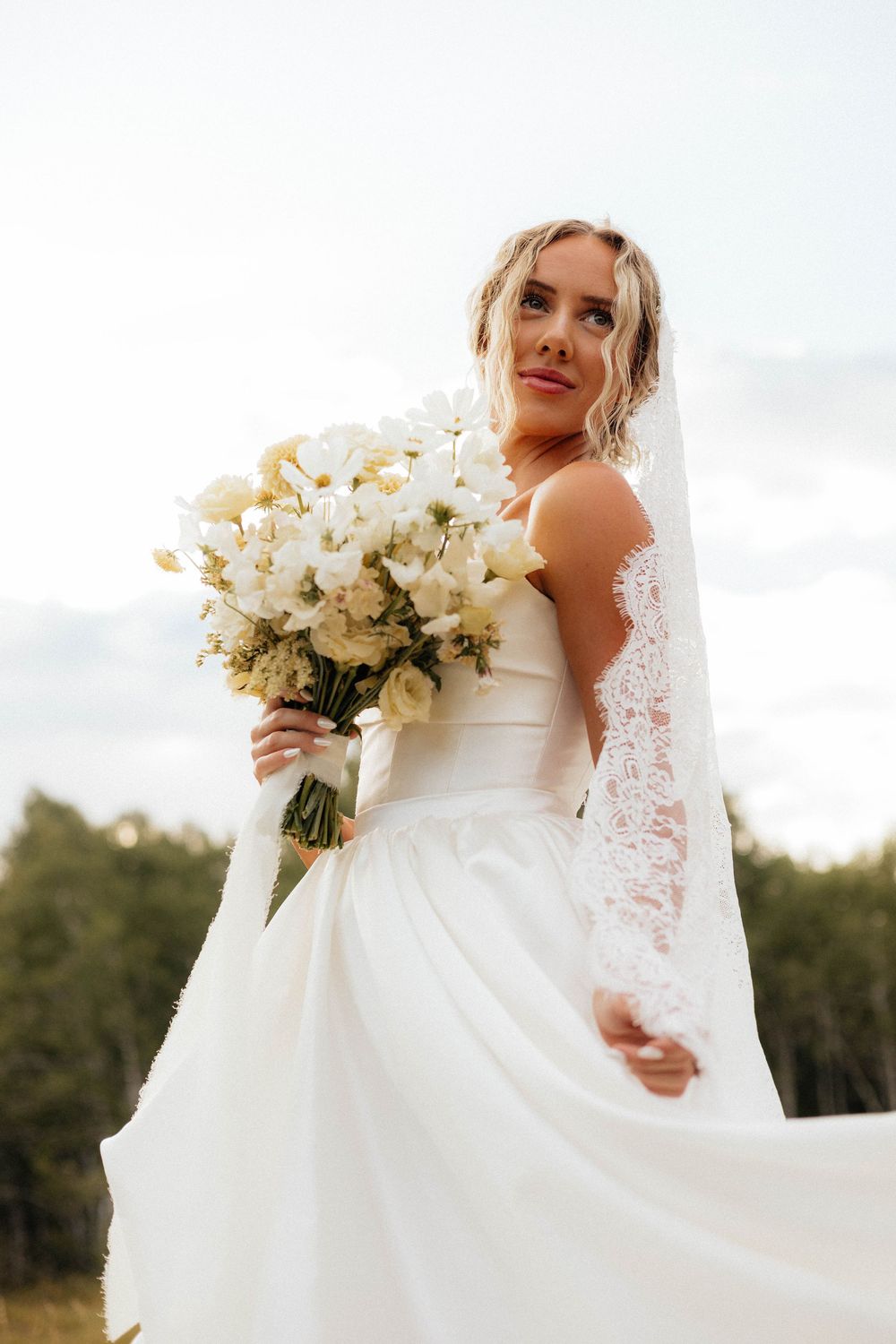 Bride in white lace gown holding bouquet of white flowers outdoors.