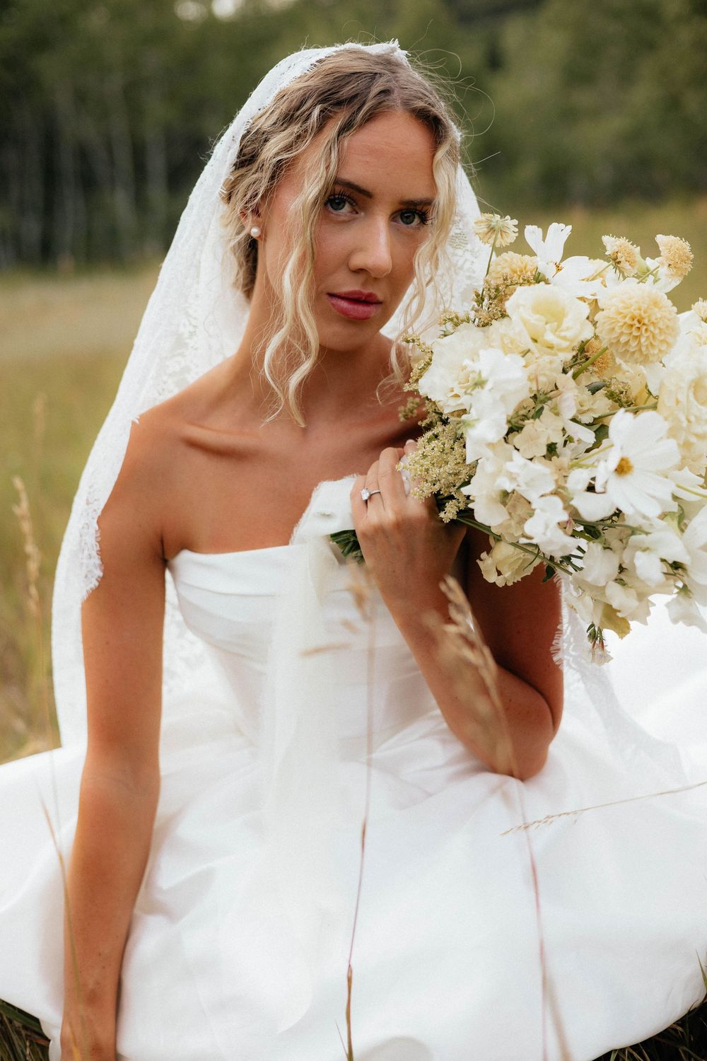 Bride in white strapless gown and veil holds white floral bouquet outdoors.