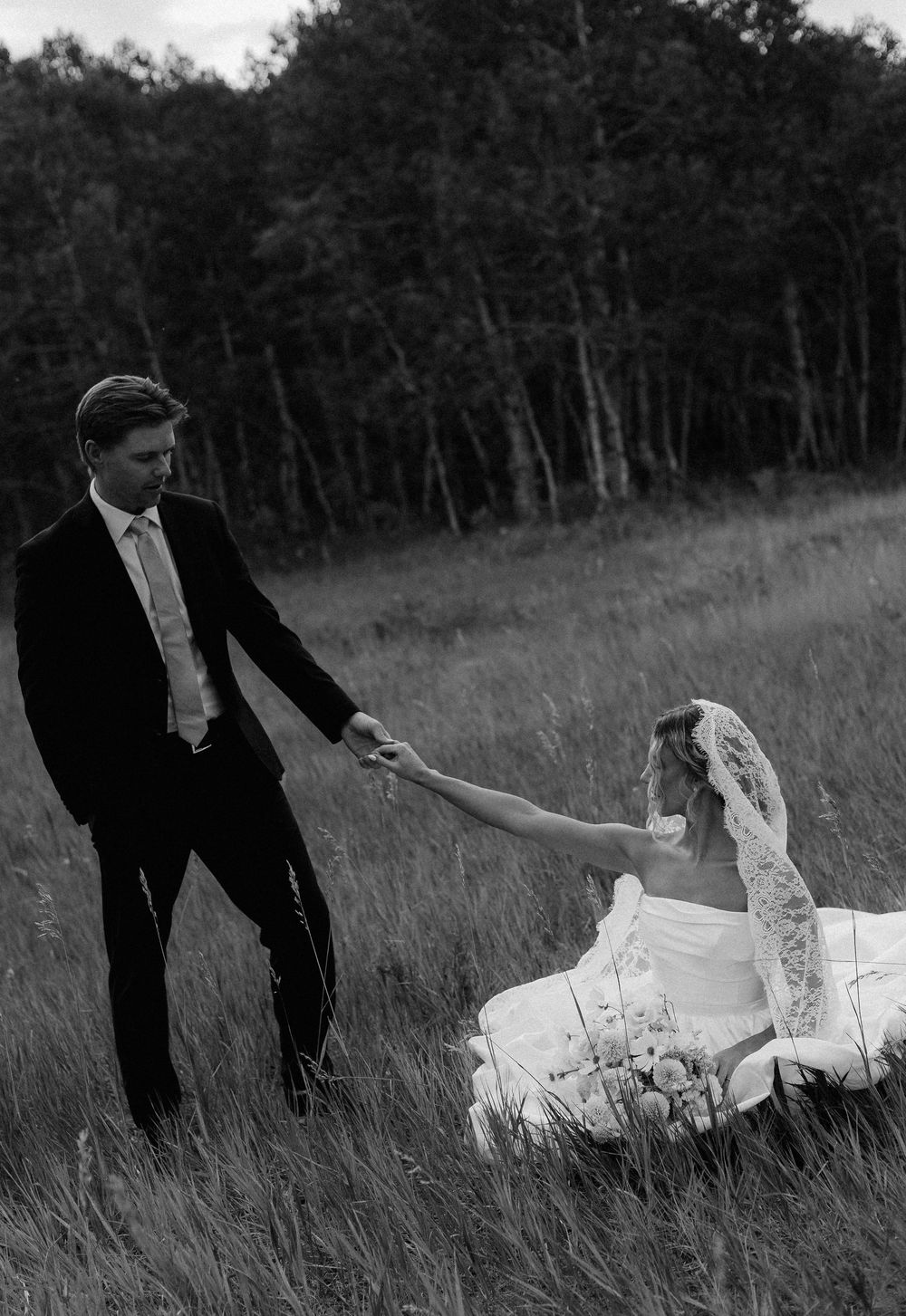 Black and white photo of groom helping bride in wedding dress rise from field of tall grass.