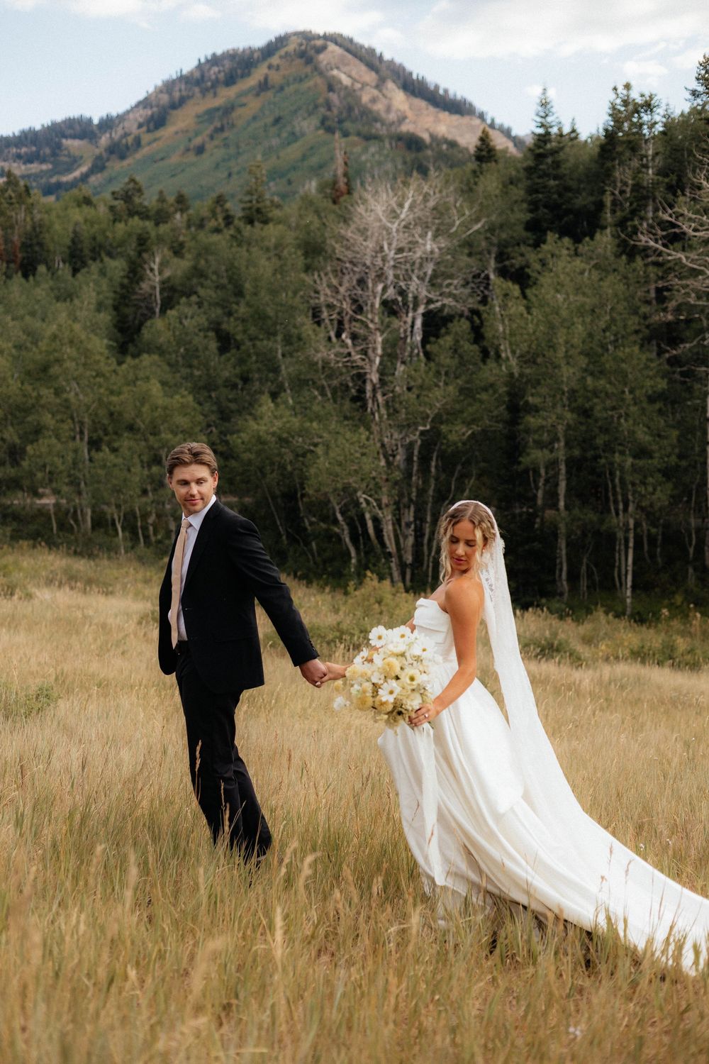 Bride and groom holding hands walking through mountain meadow, bride in flowing white gown with veil and bouquet.