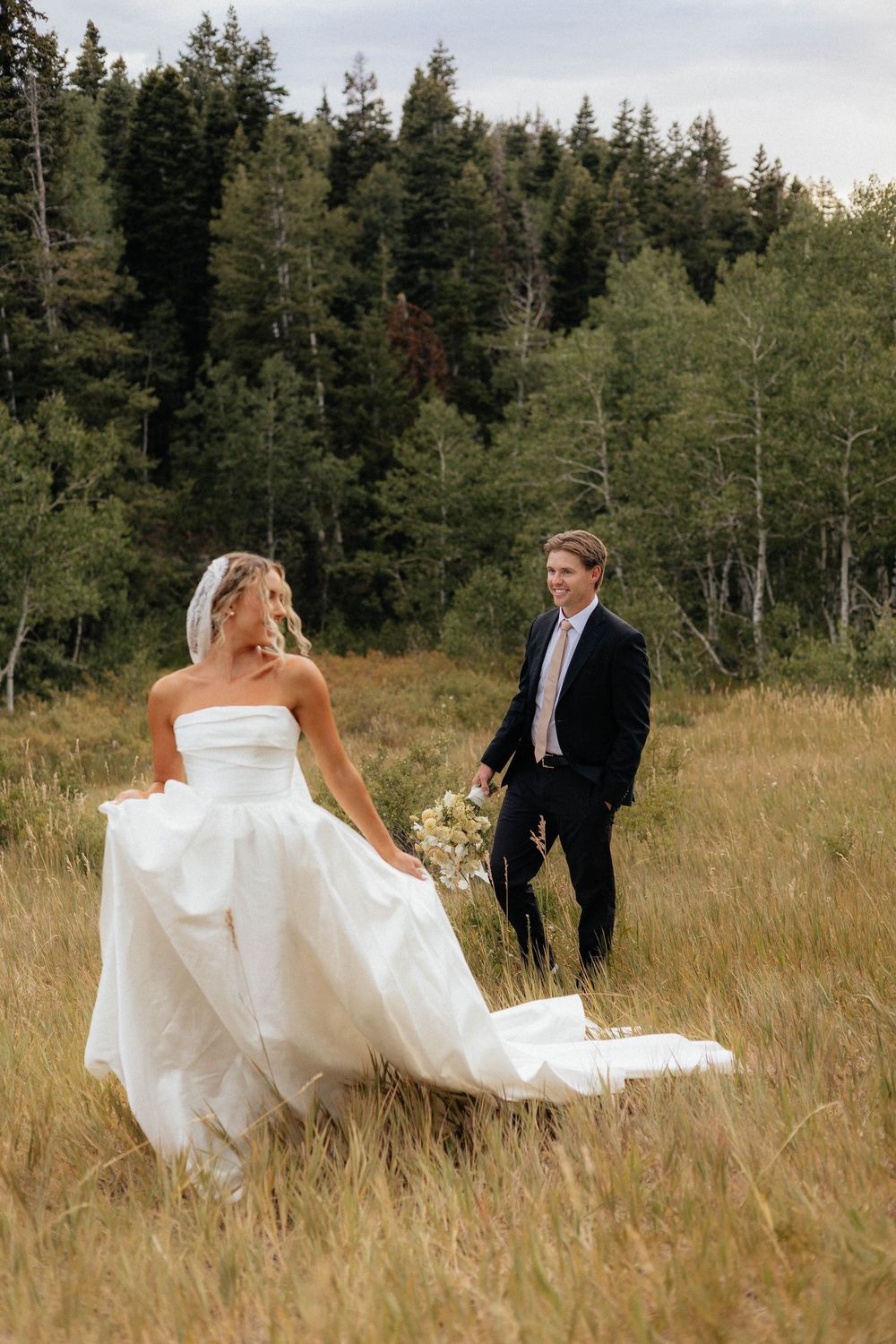Bride in white strapless ballgown walks through golden meadow as groom in black suit smiles behind her.