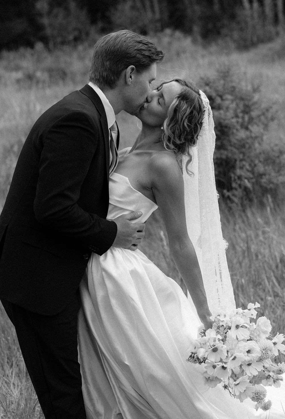 Black and white photo of a bride and groom sharing a romantic kiss outdoors, bride holding a bouquet.