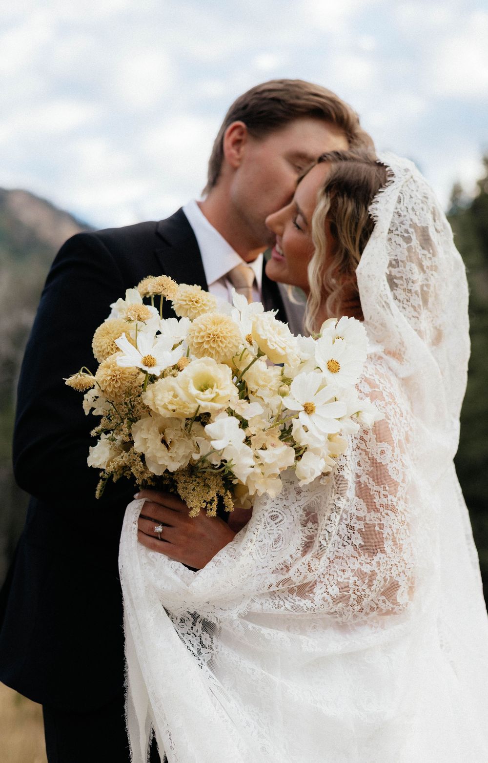 Bride and groom share tender moment, bride holding white floral bouquet with lace veil draped over shoulders.