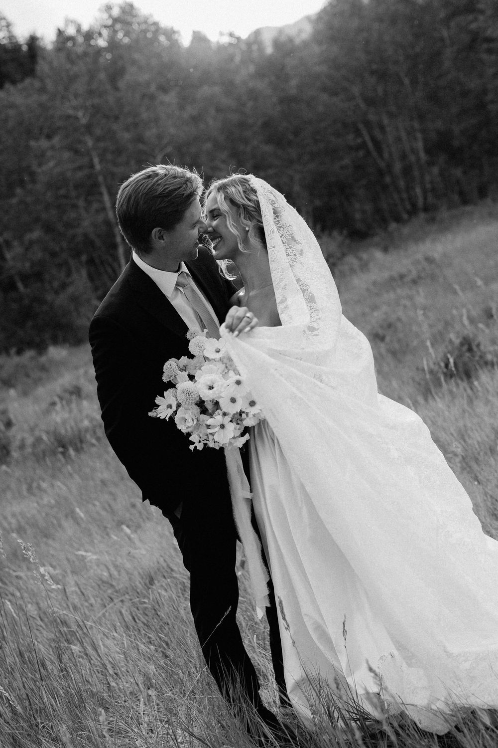 Black and white photo of bride and groom embracing in a meadow, sharing a joyful moment with bouquet in hand.