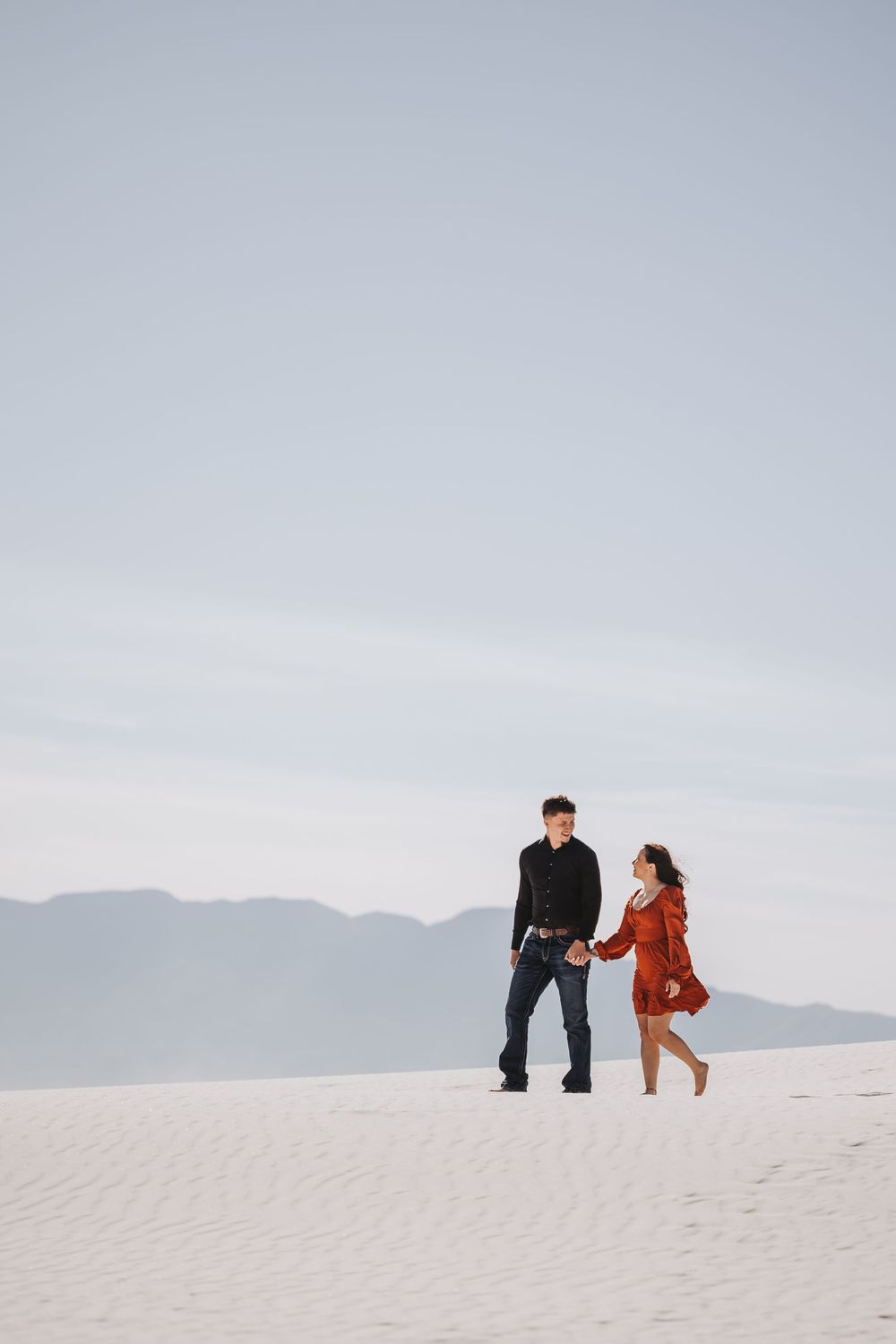 Couple holding hands walking on white sand dunes with mountain backdrop.