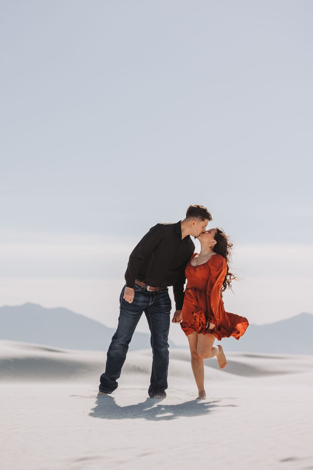 Romantic couple kissing on white sand dunes, woman in red dress, man in black.