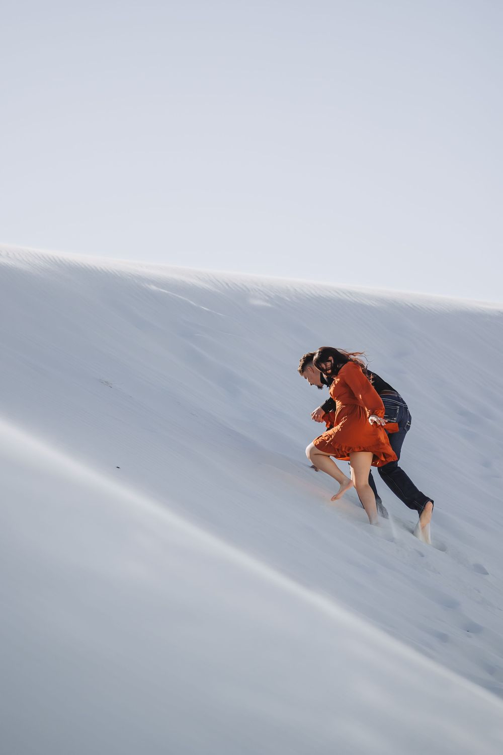 Two people climbing a white sand dune, one wearing a red dress and the other in dark clothing.
