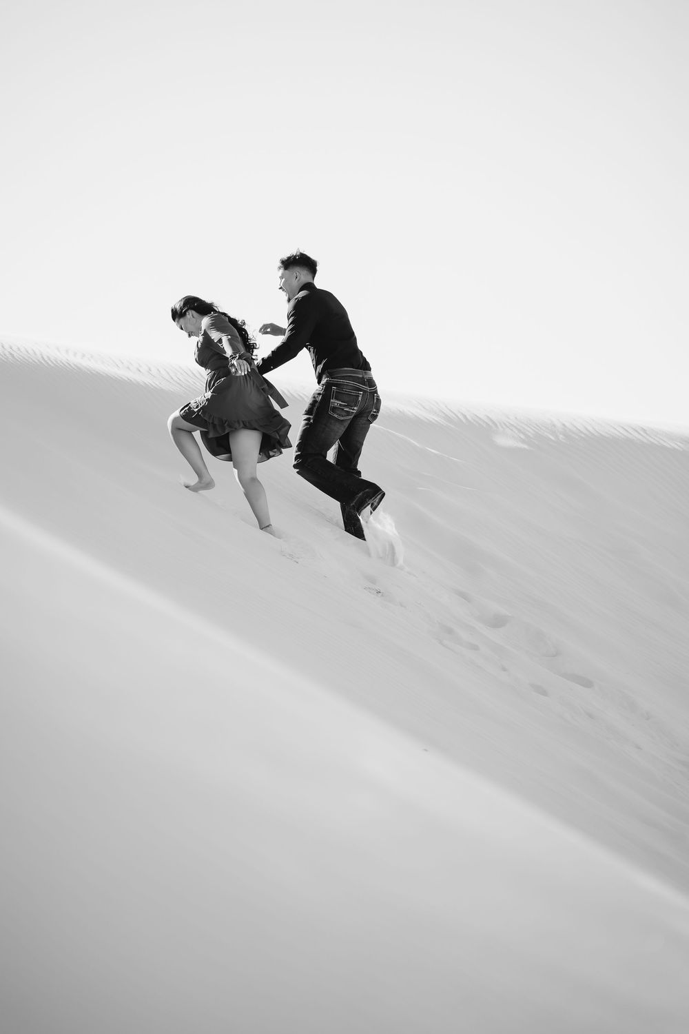 Black and white photo of a couple running and playing on a sandy desert dune, captured in motion.
