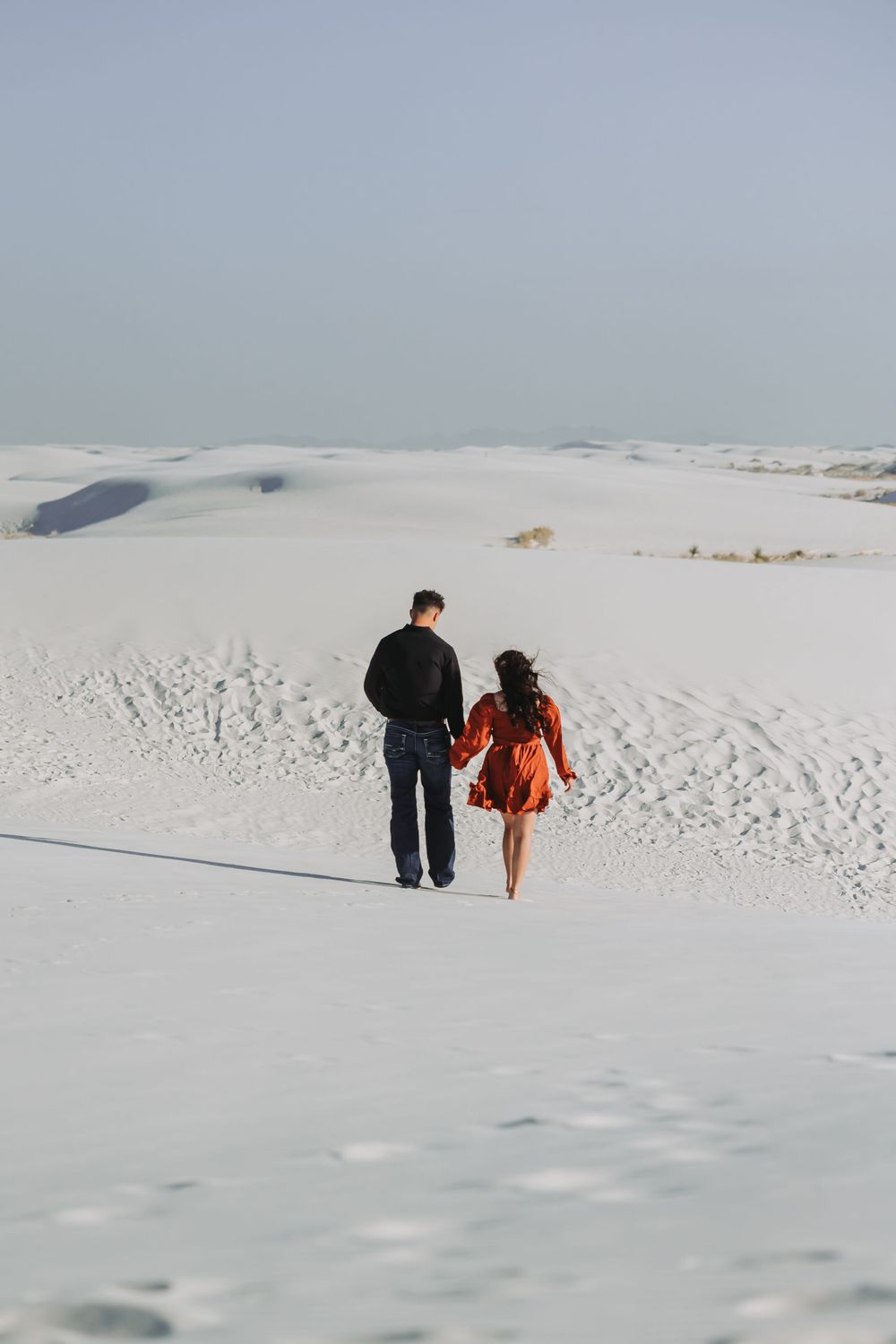 Couple holding hands walking on white sand dunes in a desert landscape, woman in orange dress and man in black jacket.