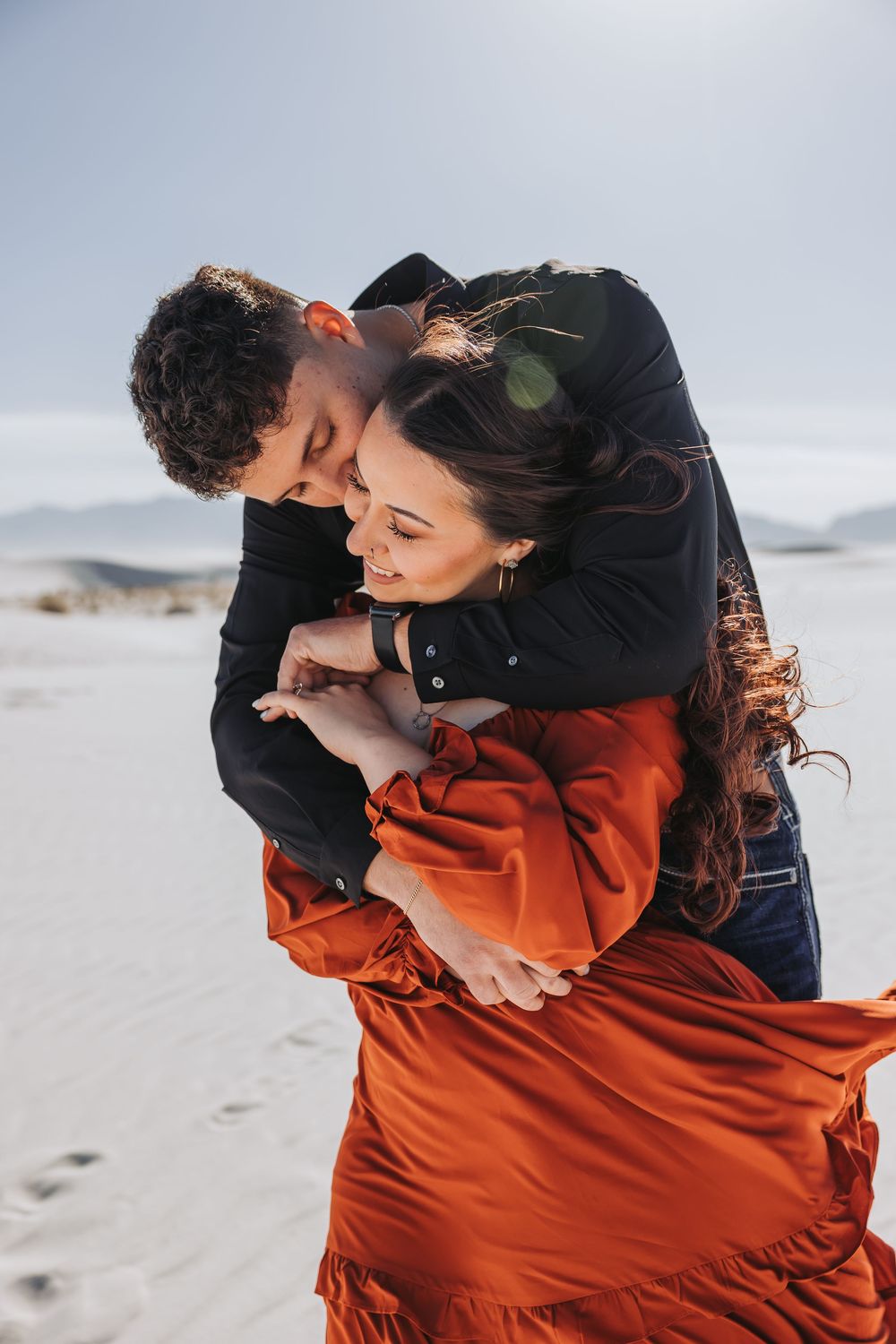 Couple embracing romantically in white sand desert, woman in flowing rust orange dress, man in black shirt.