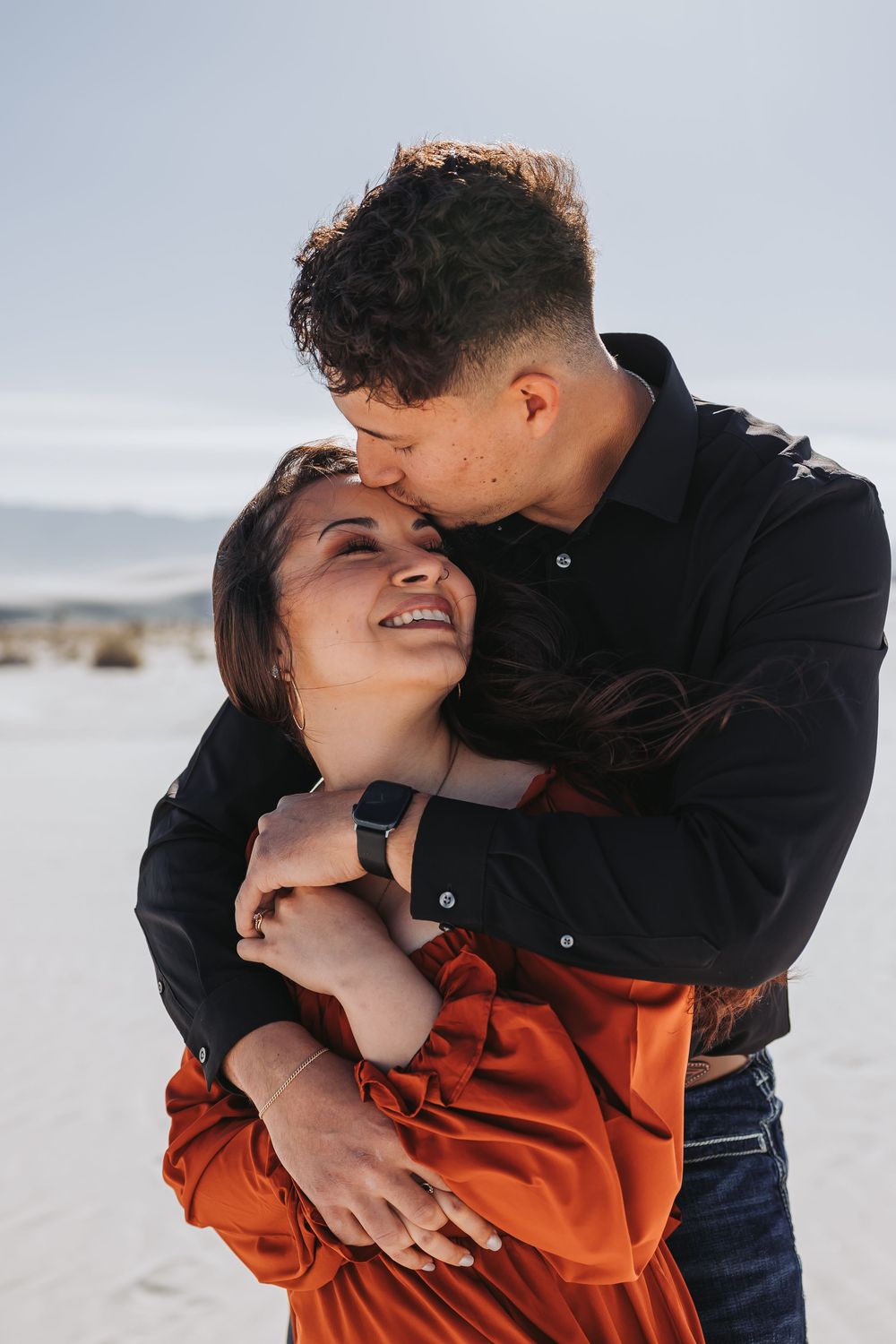 A couple embraces outdoors, man in black shirt kissing woman's head as she smiles, set against a sandy landscape.