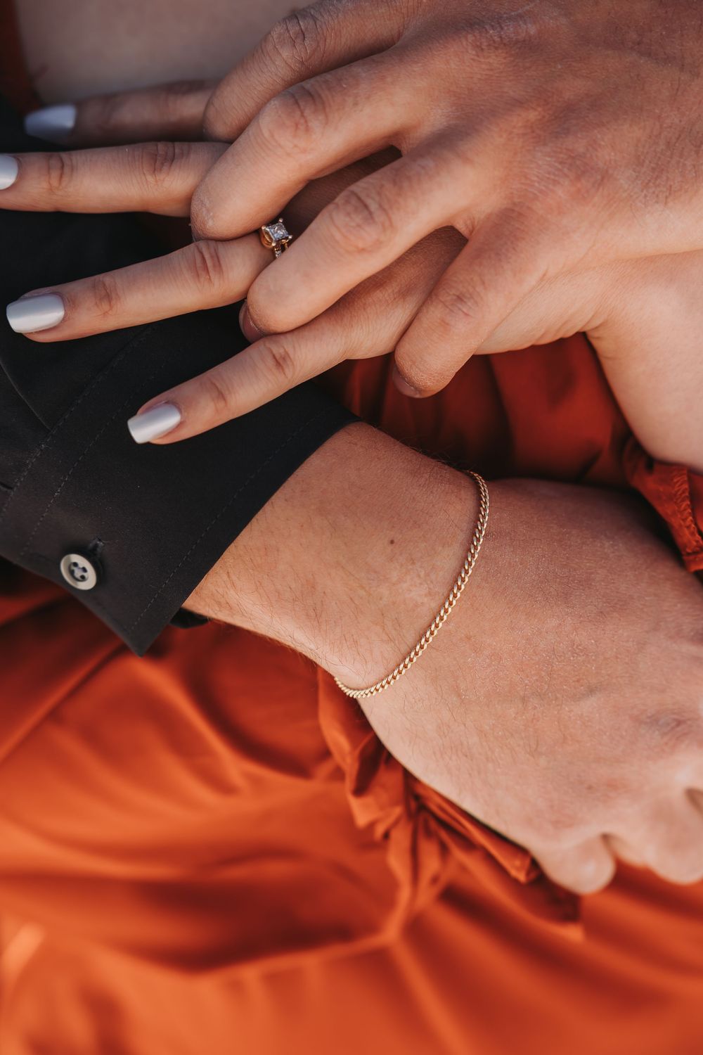 Close-up of couple's hands, woman wearing diamond engagement ring and gold chain bracelet, orange satin skirt.
