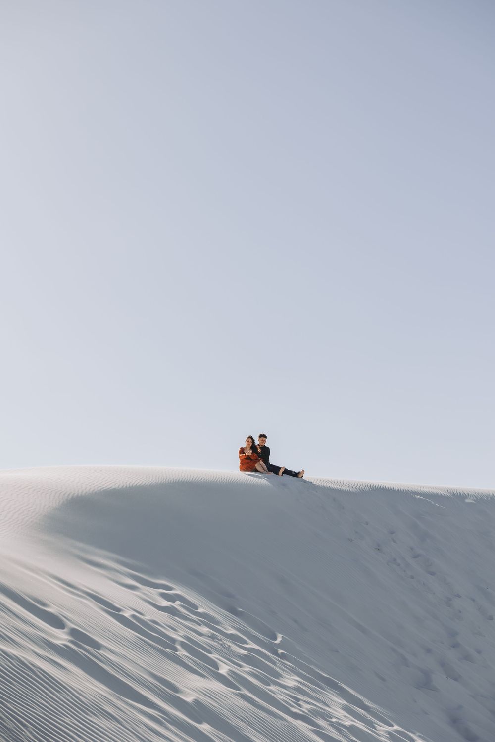 Couple sitting together on a white sand dune under a clear sky, with footprints trailing up the slope.